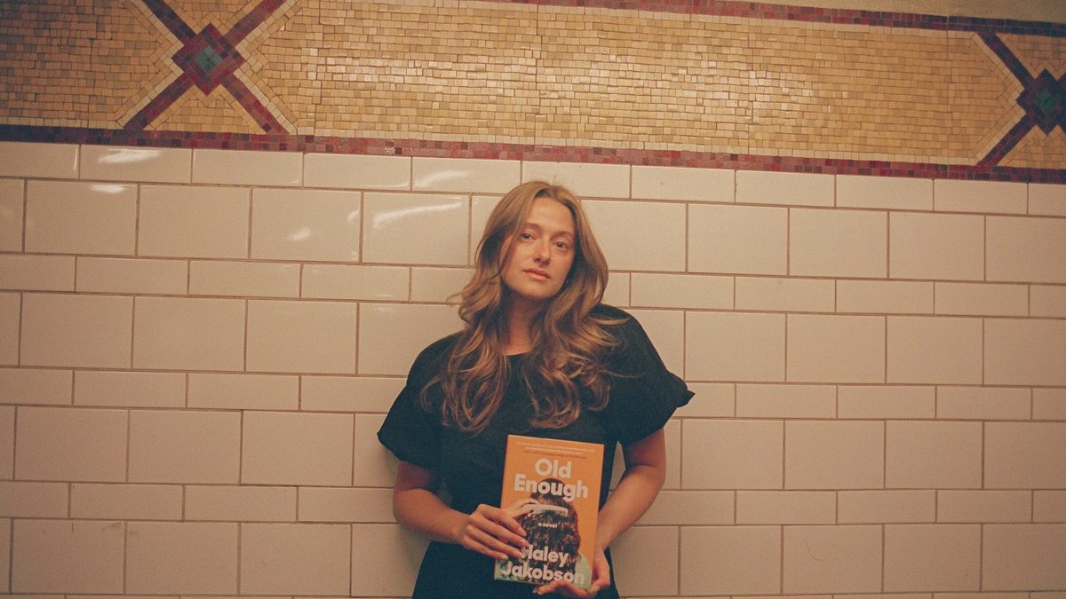 Haley Jakobson, a white queer femme, stands against an ornate tile backdrop while posing with her debut book, "Old Enough."