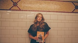 Haley Jakobson, a white queer femme, stands against an ornate tile backdrop while posing with her debut book, "Old Enough."