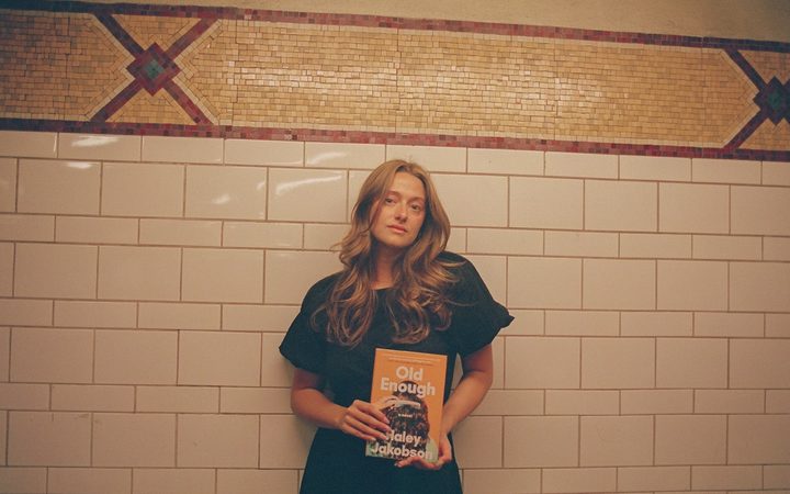 Haley Jakobson, a white queer femme, stands against an ornate tile backdrop while posing with her debut book, "Old Enough."