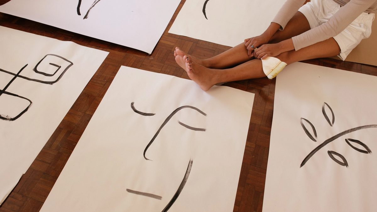A woman sits on her parquet floor, reflecting on the abstract line art she has painted on posters laid out before her.