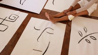 A woman sits on her parquet floor, reflecting on the abstract line art she has painted on posters laid out before her.