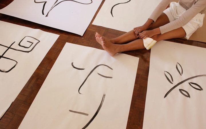 A woman sits on her parquet floor, reflecting on the abstract line art she has painted on posters laid out before her.