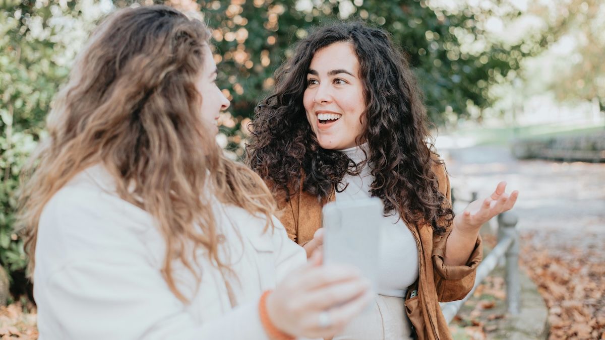 Two women sitting in a park, looking surprised by something on their phone