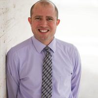 Gary, a sharply dressed therapist in a lilac oxford shirt and tie, smiling to camera in a brightly-lit room.