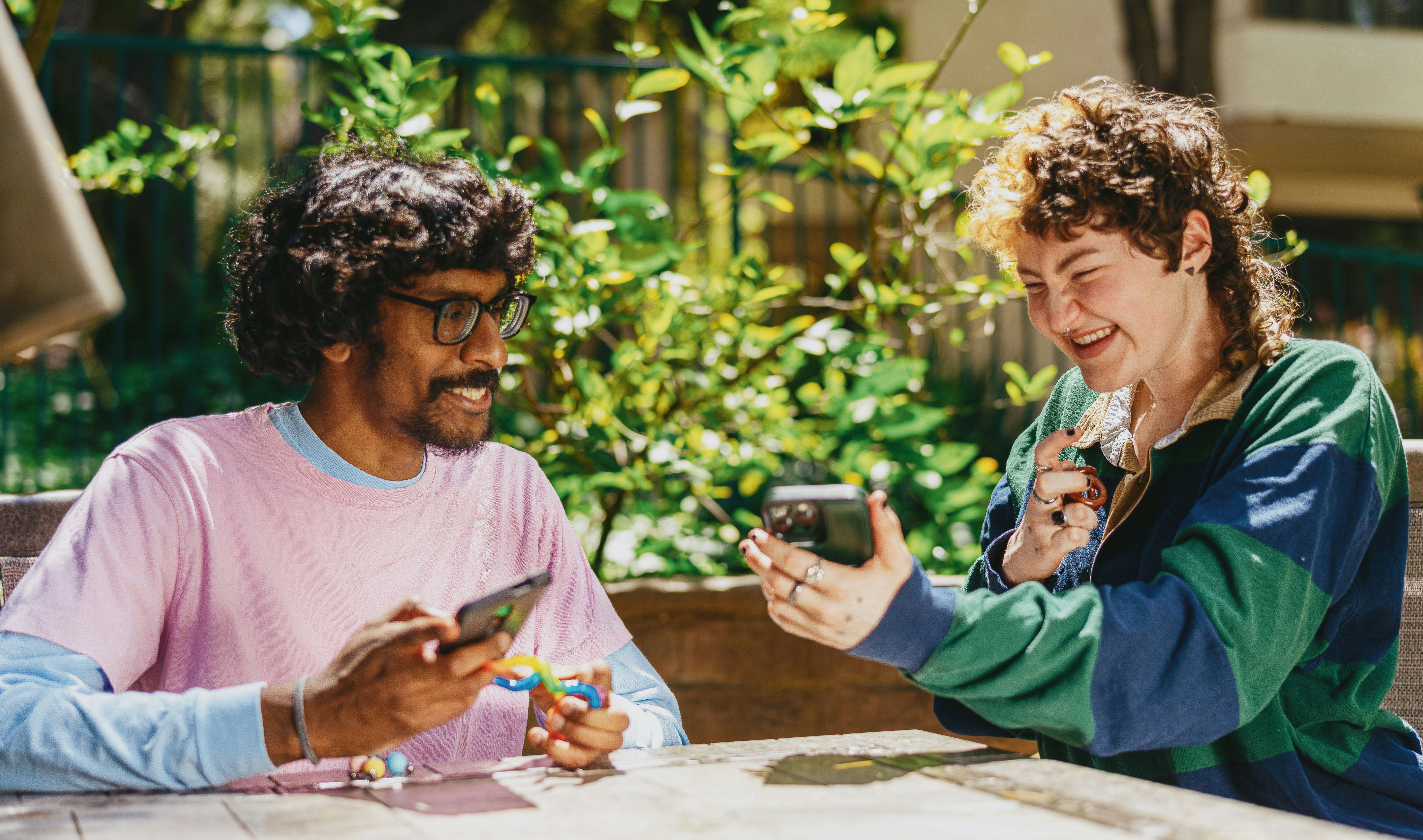 Two individuals sit outside, surrounded by greenery. They are both smiling, holding fidget toys in one hand and a cell phone in another.