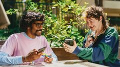 Two individuals sit outside, surrounded by greenery. They are both smiling, holding fidget toys in one hand and a cell phone in another.