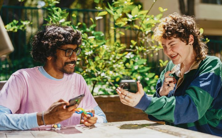 Two individuals sit outside, surrounded by greenery. They are both smiling, holding fidget toys in one hand and a cell phone in another.
