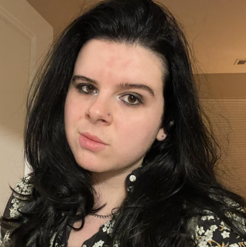 Adryan, a white nonbinary journalist with long brown hair stares into the camera, with an eyebrow raised and a slight grin.