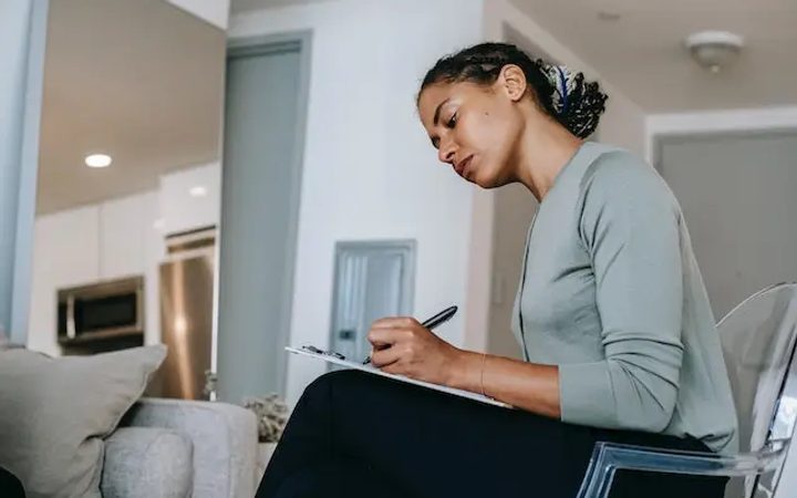 A Black therapist taking progress notes on her clipboard while seated in front of a patient.