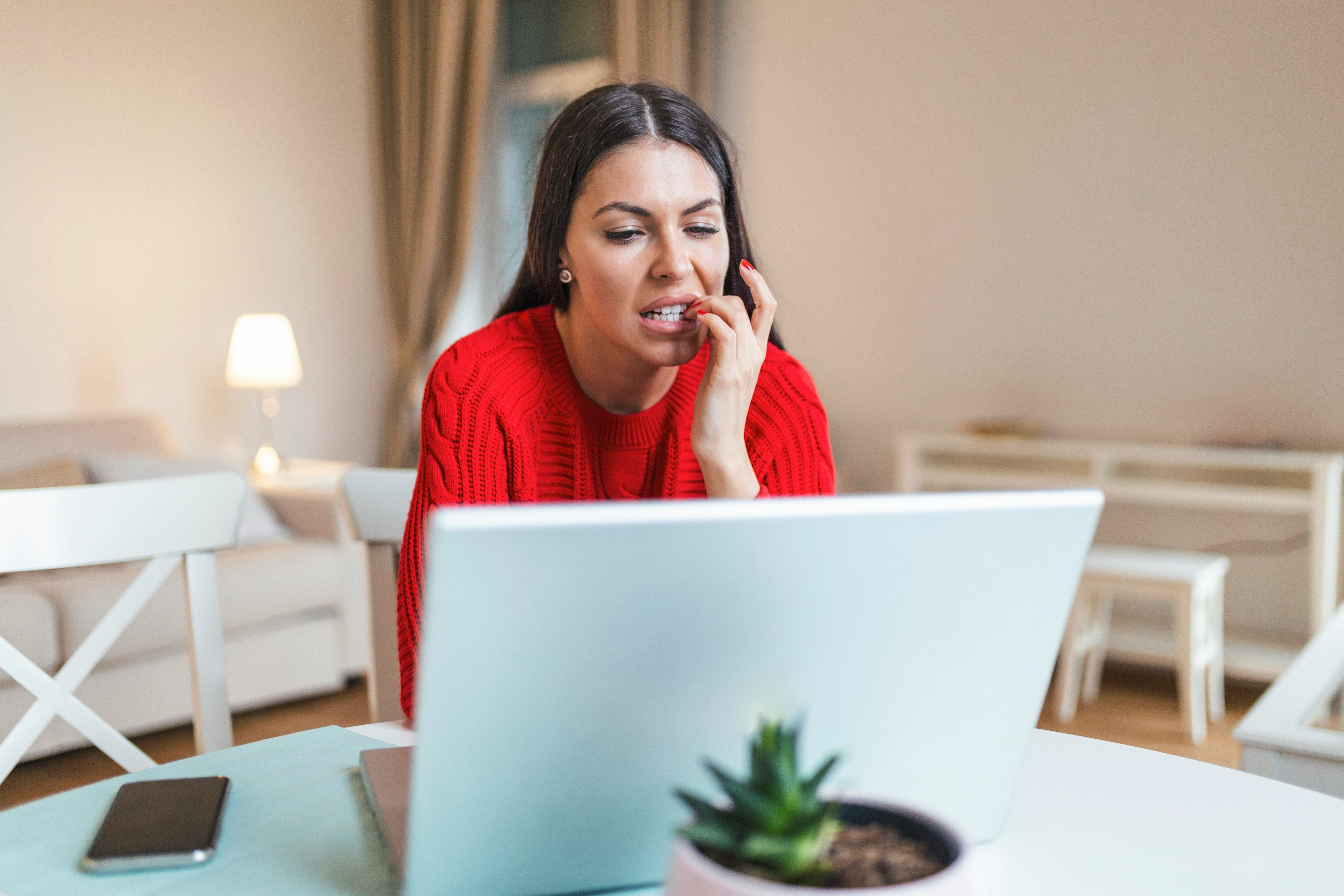 Woman in a red sweater looking anxious while working on a laptop at home.