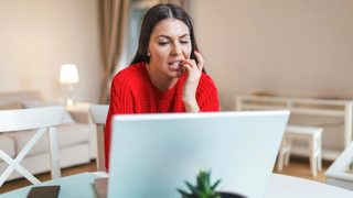 Woman in a red sweater looking anxious while working on a laptop at home.