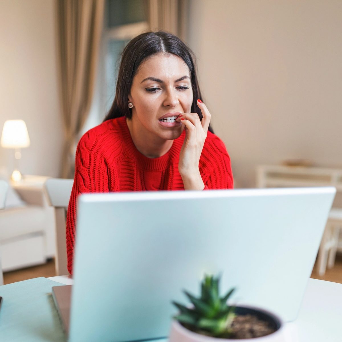 Woman in a red sweater looking anxious while working on a laptop at home.