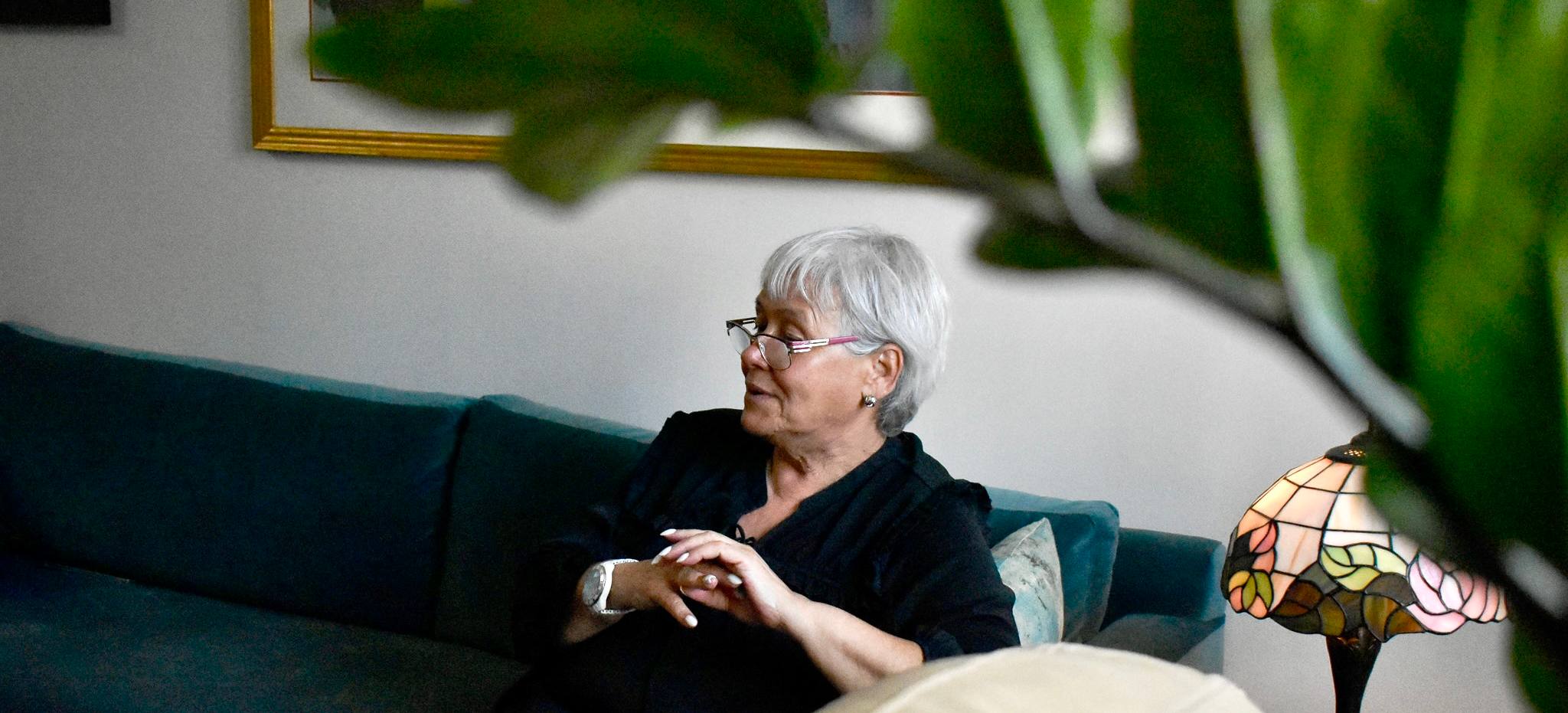 An older woman with short grey hair sitting on a sofa, looking toward a window