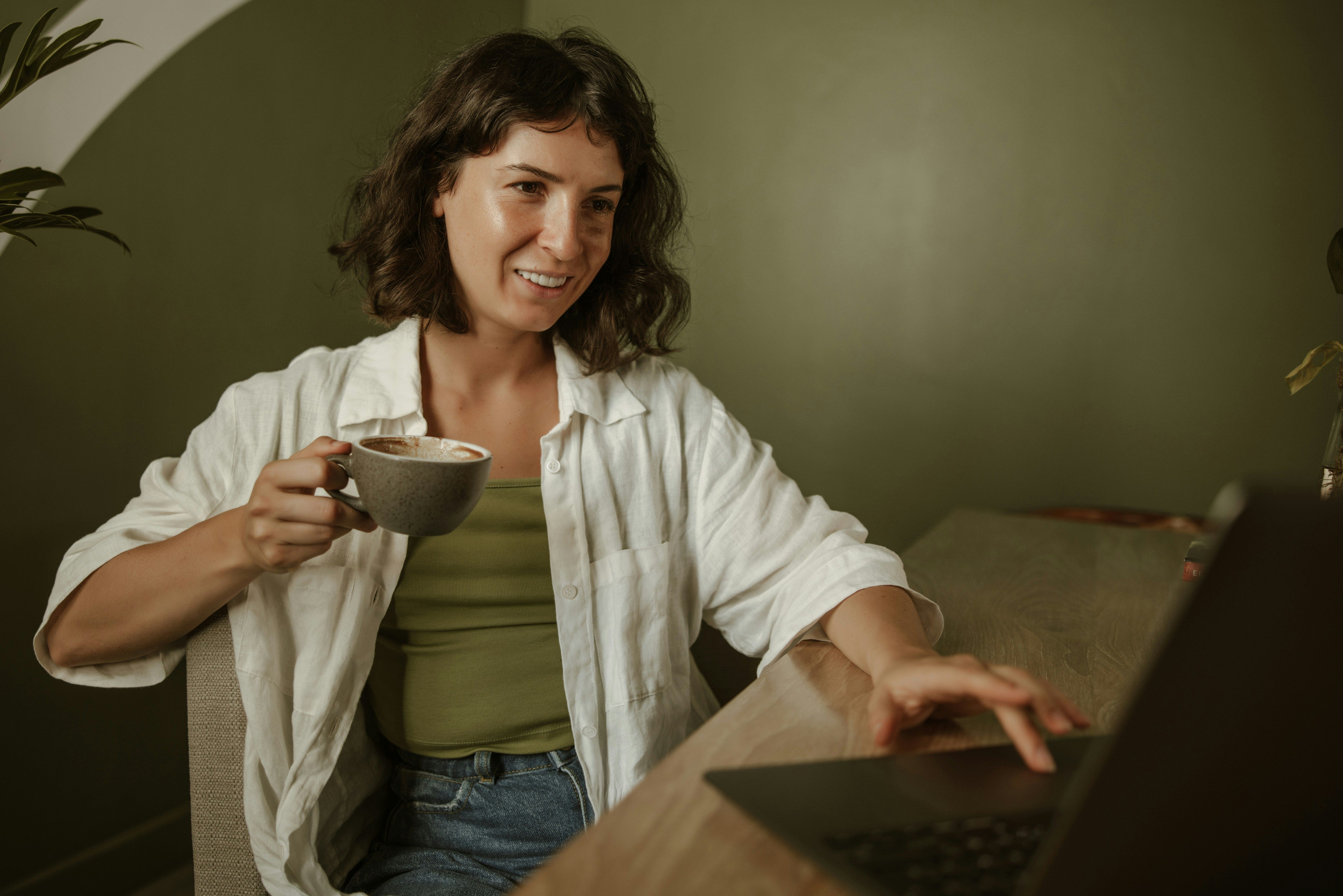 Woman sitting at a desk smiling and holding a coffee cup while on a laptop