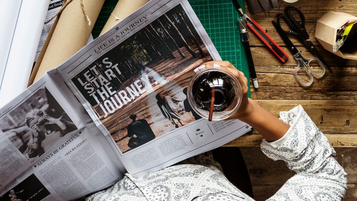 A woman is at her desk drinking coffee and getting ready to create her vision board, starting with a newspaper cutout that reads "let's start the journey"