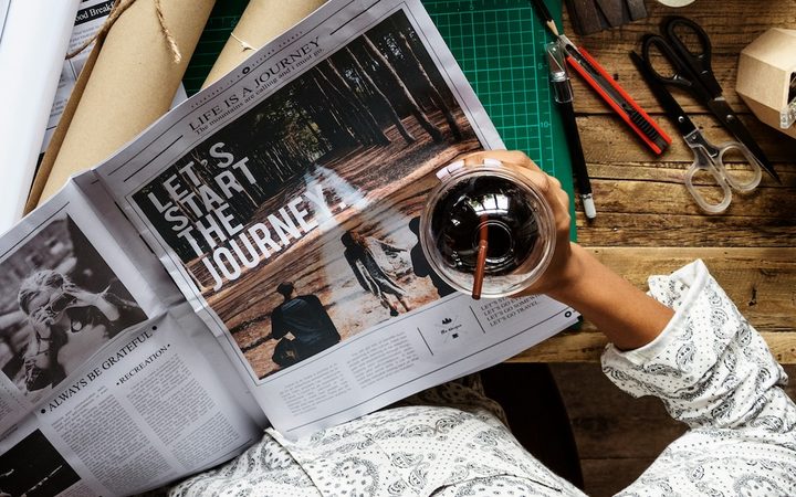 A woman is at her desk drinking coffee and getting ready to create her vision board, starting with a newspaper cutout that reads "let's start the journey"