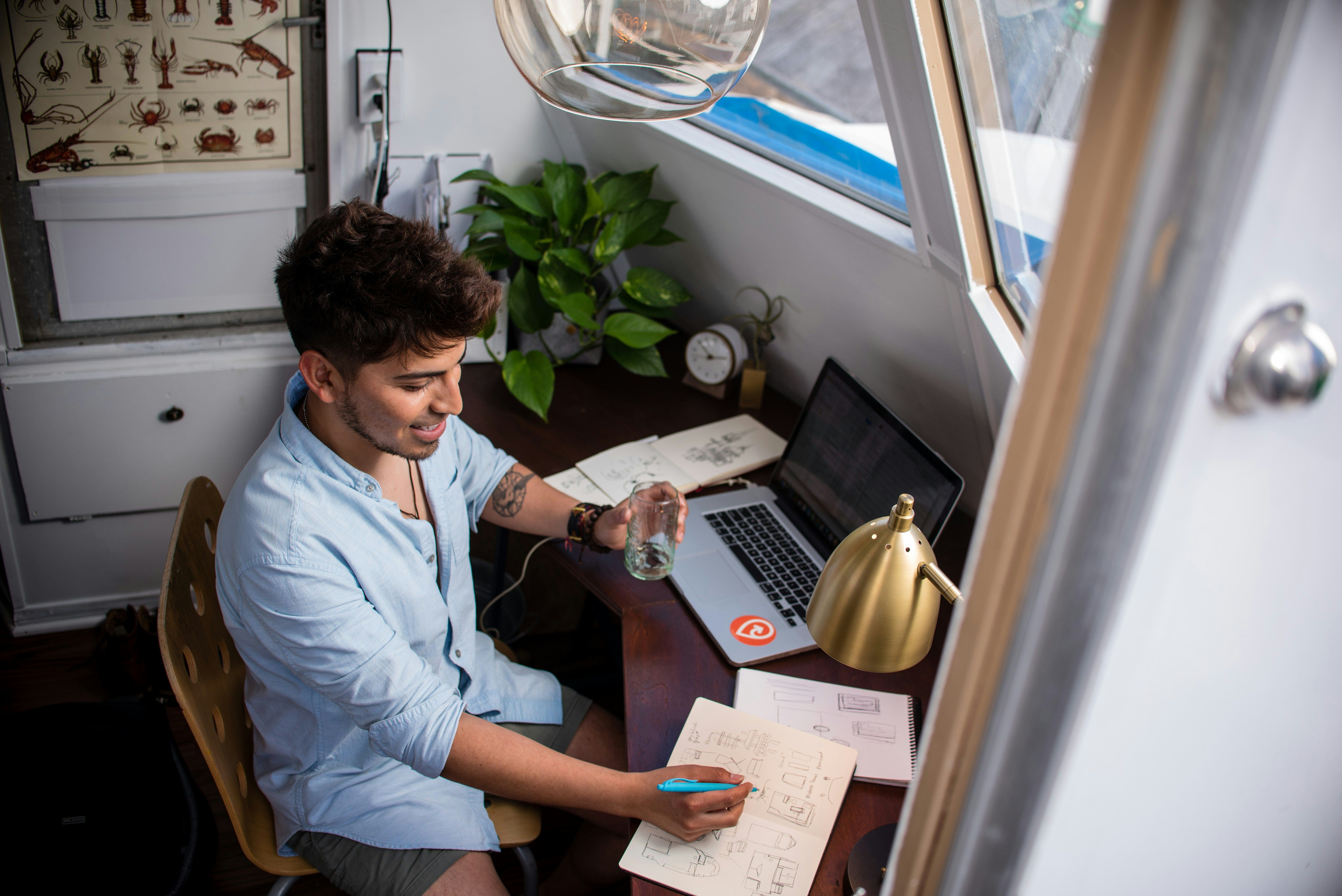 Someone in a baby blue button-up and short hair sits at a cluttered desk, glass of water in hand, marking up a notebook at their desk. Scattered across the desk is a laptop, lamp, additional papers, and a plant. They appear cheerful but busy.