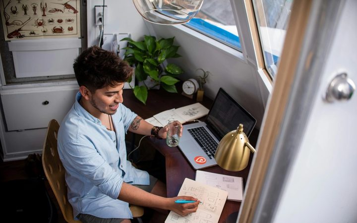 Someone in a baby blue button-up and short hair sits at a cluttered desk, glass of water in hand, marking up a notebook at their desk. Scattered across the desk is a laptop, lamp, additional papers, and a plant. They appear cheerful but busy.