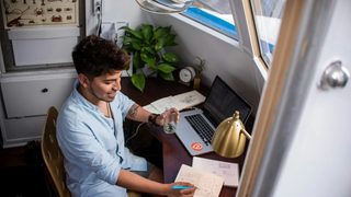 Someone in a baby blue button-up and short hair sits at a cluttered desk, glass of water in hand, marking up a notebook at their desk. Scattered across the desk is a laptop, lamp, additional papers, and a plant. They appear cheerful but busy.