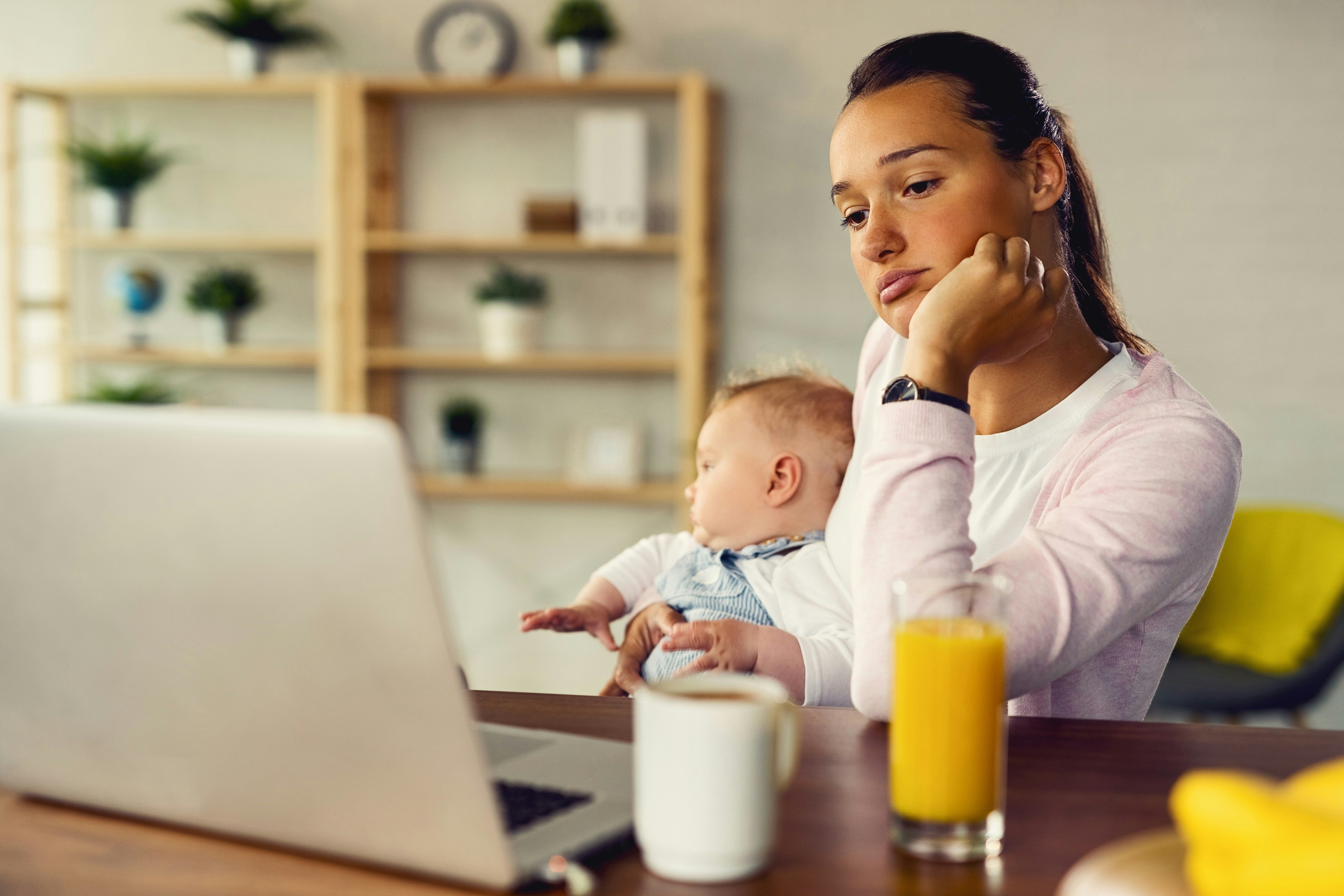 Mother sits at desk holding her baby and looking at her laptop