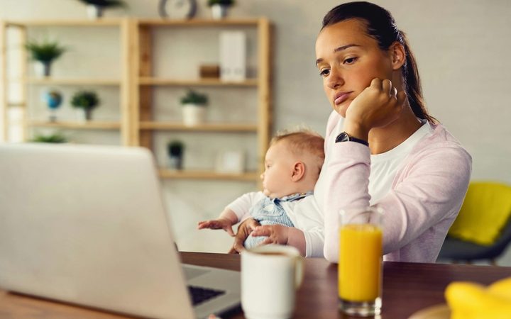 Mother sits at desk holding her baby and looking at her laptop