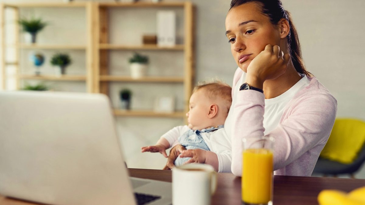 Mother sits at desk holding her baby and looking at her laptop
