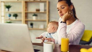 Mother sits at desk holding her baby and looking at her laptop