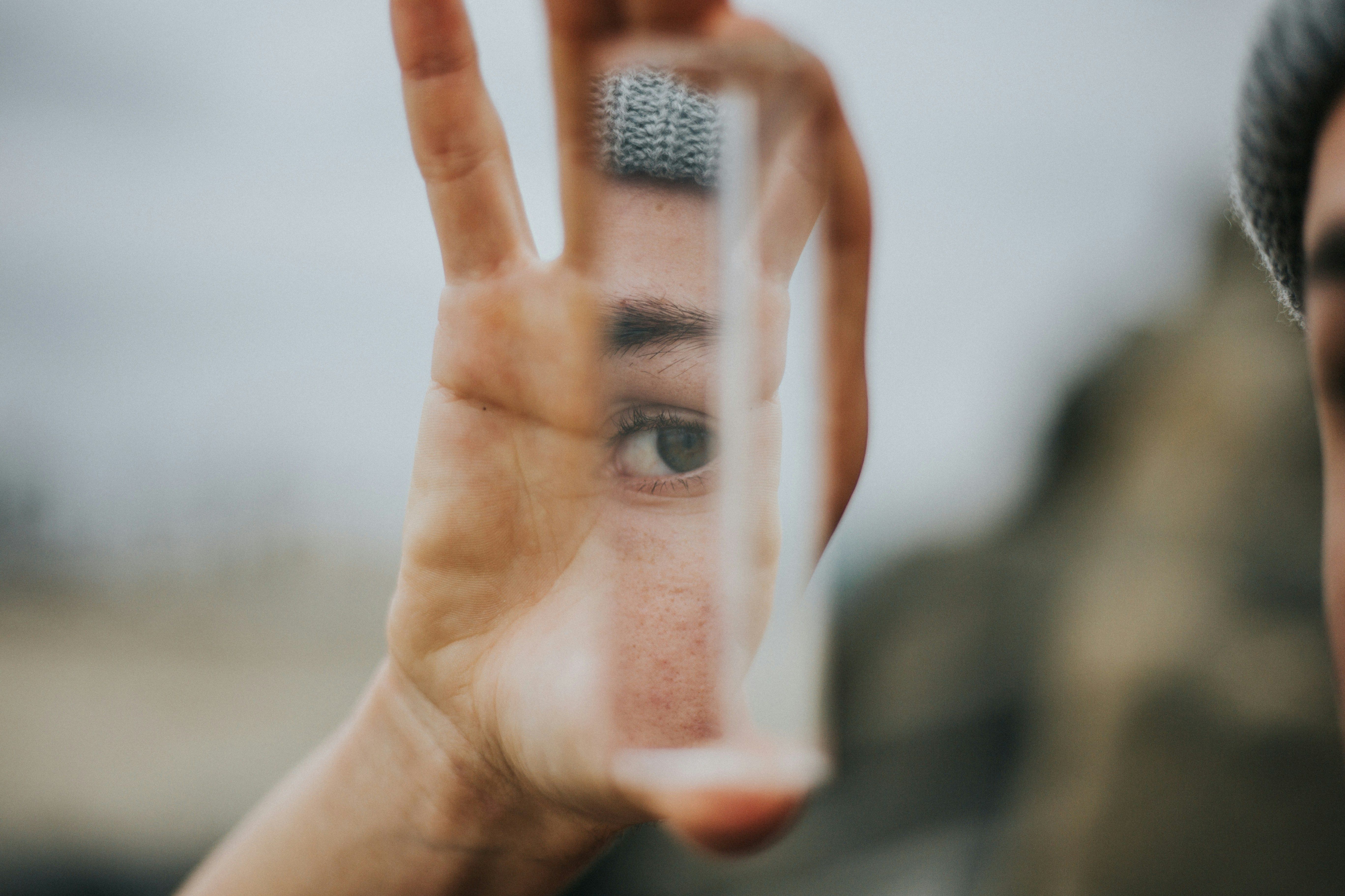 A hand holds up a reflective glass fragment, revealing a small sliver of someone's face, including a pensive eye staring back.