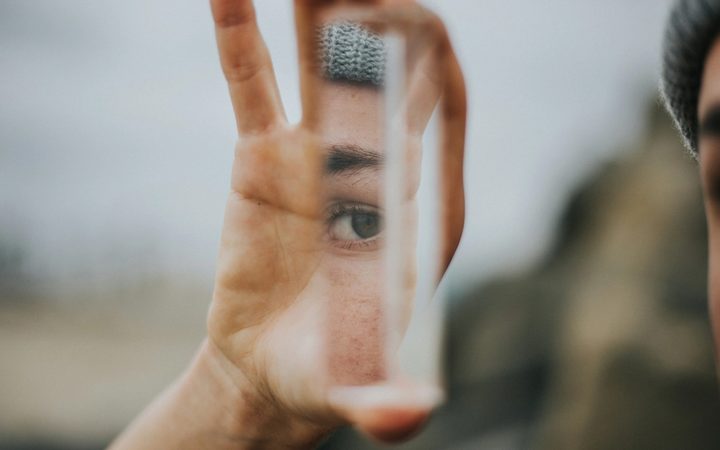 A hand holds up a reflective glass fragment, revealing a small sliver of someone's face, including a pensive eye staring back.