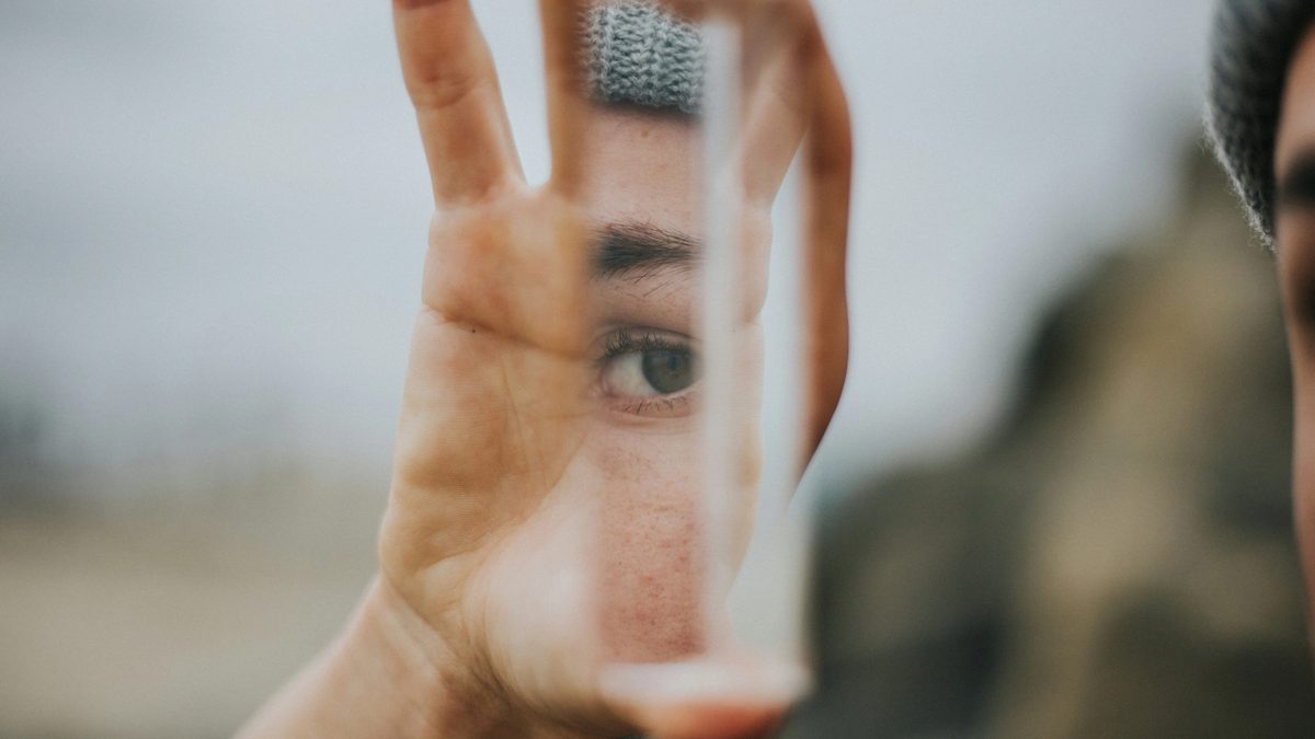 A hand holds up a reflective glass fragment, revealing a small sliver of someone's face, including a pensive eye staring back.