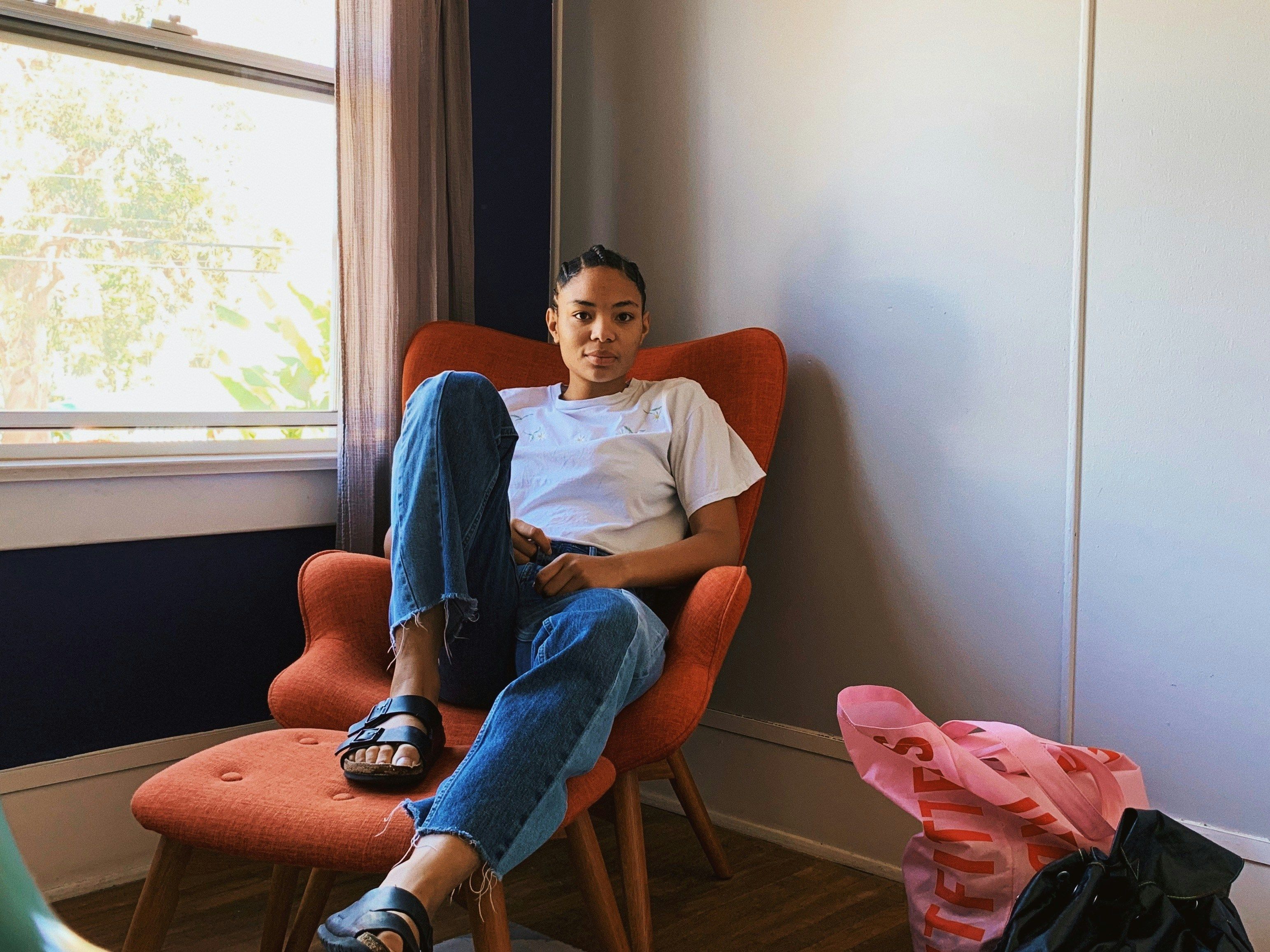 A person with dark skin and braids pulled back sits in a relaxed, reclined position on a bright orange armchair. One foot is flat on the ottoman in front of them, while the other dangles off with their leg stretched out. They are seated by a bright window in the corner of a room.