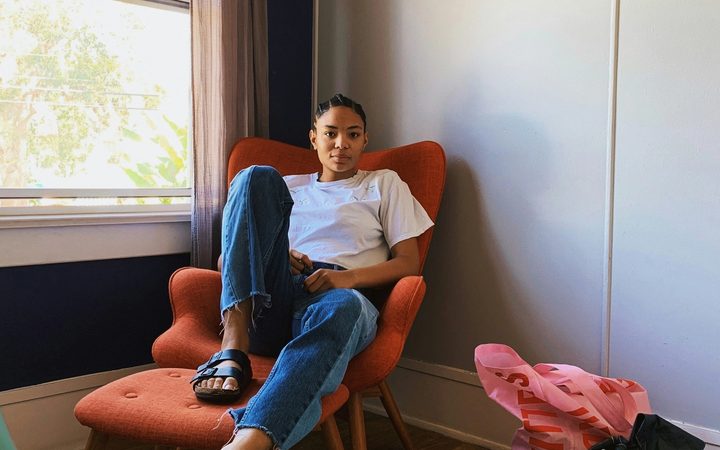 A person with dark skin and braids pulled back sits in a relaxed, reclined position on a bright orange armchair. One foot is flat on the ottoman in front of them, while the other dangles off with their leg stretched out. They are seated by a bright window in the corner of a room.