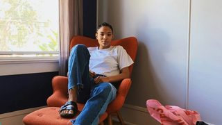 A person with dark skin and braids pulled back sits in a relaxed, reclined position on a bright orange armchair. One foot is flat on the ottoman in front of them, while the other dangles off with their leg stretched out. They are seated by a bright window in the corner of a room.