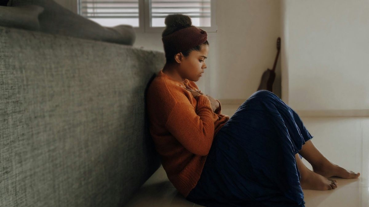Overcome by a wave of grief, a young woman is sitting on the floor behind a couch, covering her heart with her hands