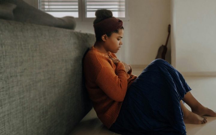 Overcome by a wave of grief, a young woman is sitting on the floor behind a couch, covering her heart with her hands