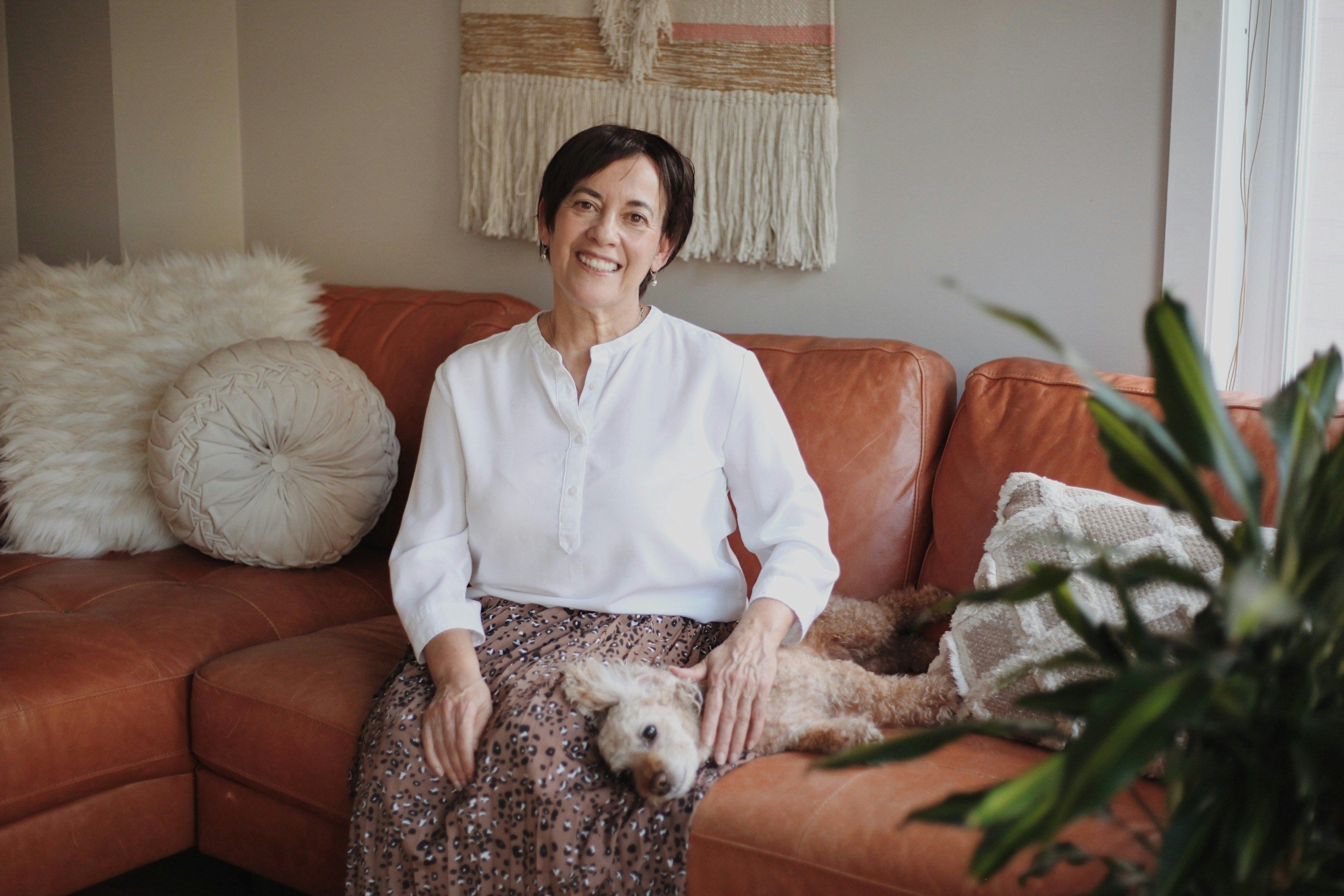 A feminine-presenting elder with short, dark hair sits on a couch, smiling and gazing into the camera as a small, white dog reclines across their lap. The couch they're seated upon is a stylish rust color, with decorative pillows, a plant, and a macrame wall hanging displayed above them.