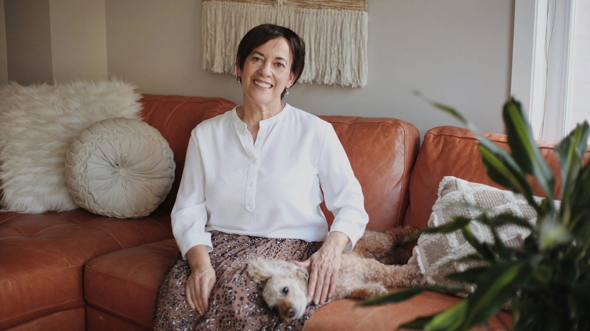 A feminine-presenting elder with short, dark hair sits on a couch, smiling and gazing into the camera as a small, white dog reclines across their lap. The couch they're seated upon is a stylish rust color, with decorative pillows, a plant, and a macrame wall hanging displayed above them.