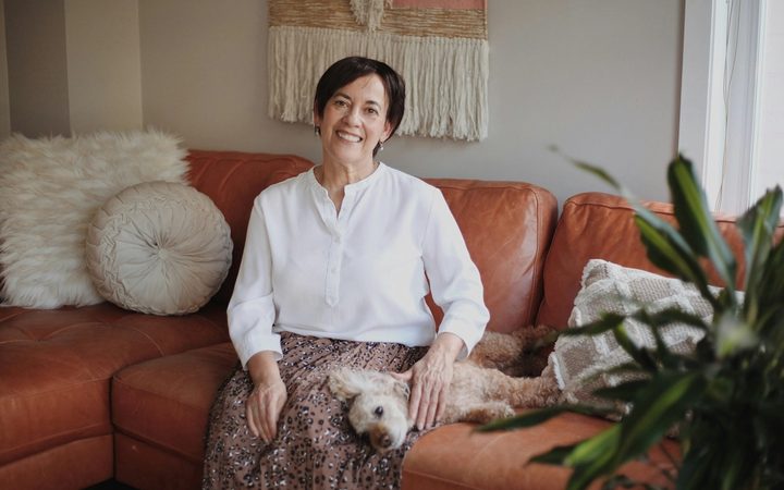 A feminine-presenting elder with short, dark hair sits on a couch, smiling and gazing into the camera as a small, white dog reclines across their lap. The couch they're seated upon is a stylish rust color, with decorative pillows, a plant, and a macrame wall hanging displayed above them.
