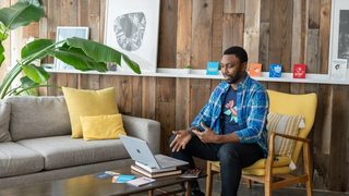 Man in blue and white plaid dress shirt and black pants sitting on couch using macbook