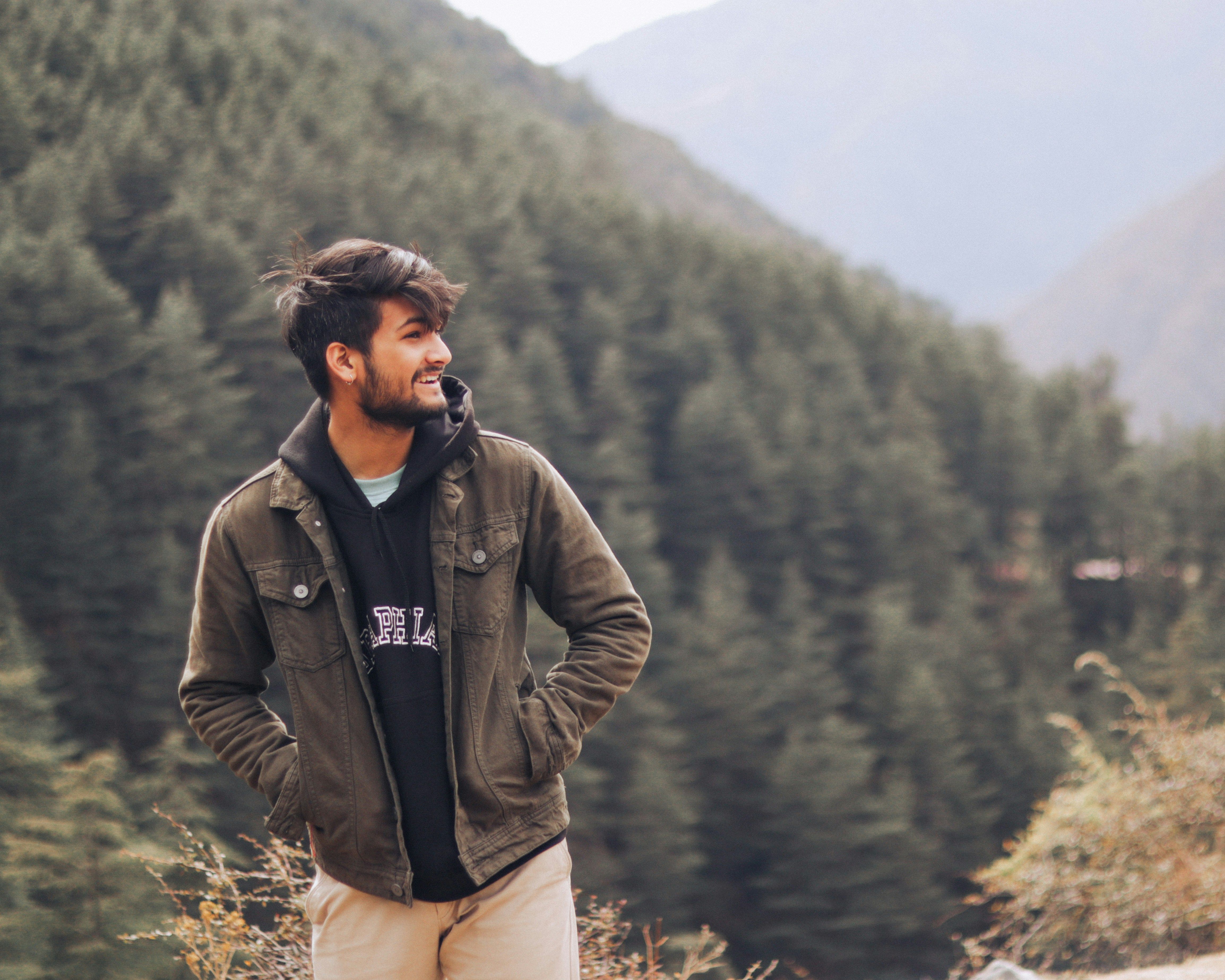 Man smiling while standing outdoors in a forested mountain landscape, wearing a jacket and hoodie with hands in pockets.