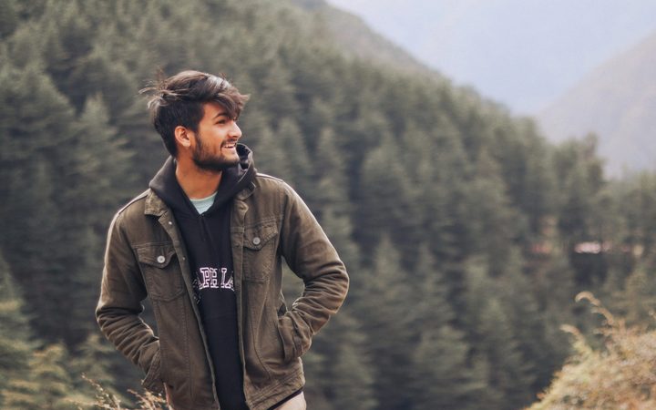 Man smiling while standing outdoors in a forested mountain landscape, wearing a jacket and hoodie with hands in pockets.