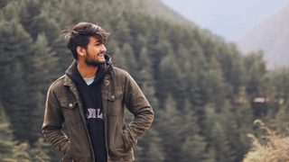 Man smiling while standing outdoors in a forested mountain landscape, wearing a jacket and hoodie with hands in pockets.