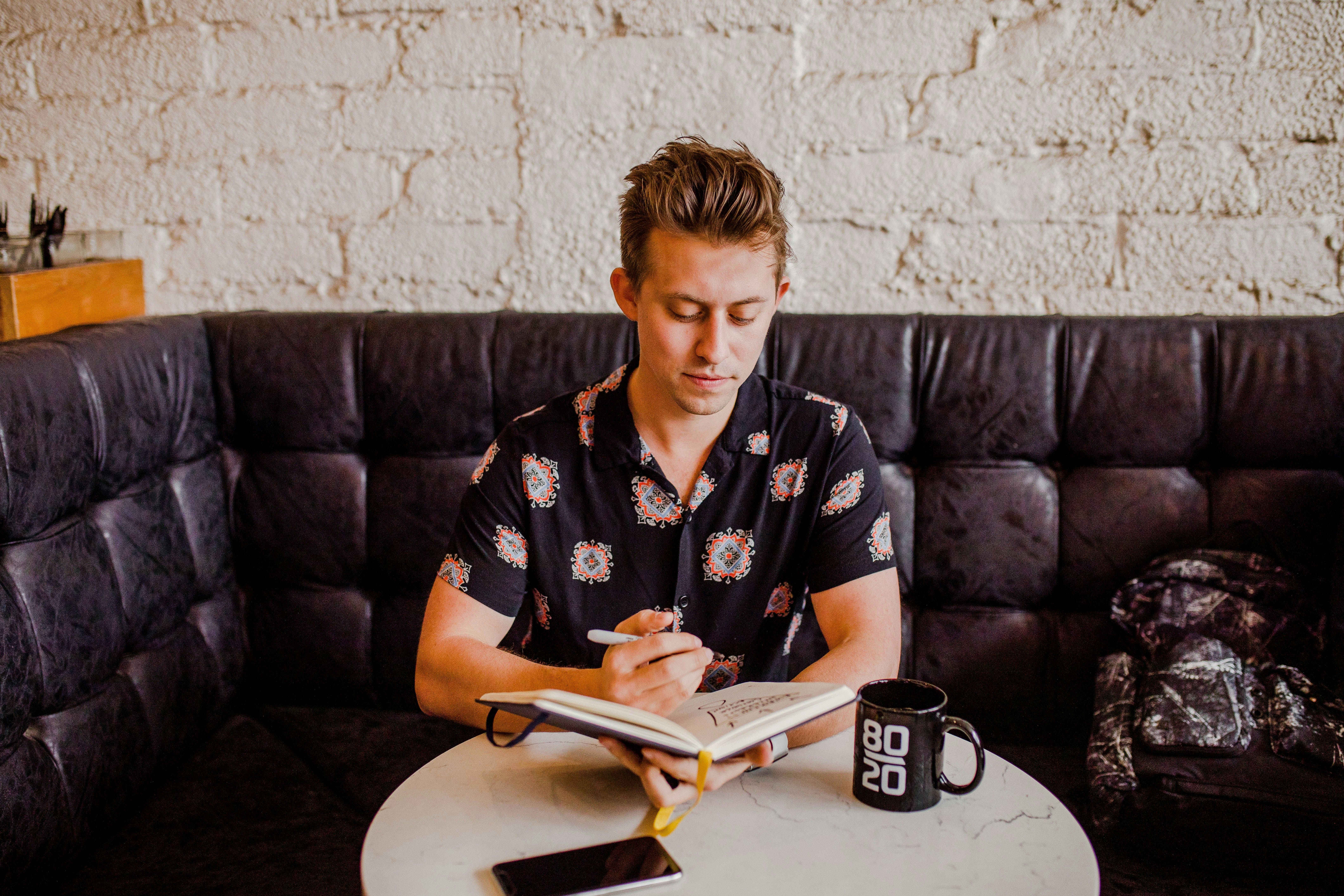 A masculine-appearing person with short hair and a patterned button-up sits in a leather booth at a cafe, looking down at an open notebook with a coffee cup and smartphone within reach. They appear lost in thought, pen in hand, as if contemplating something to write.