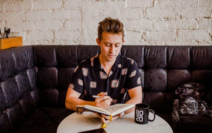A masculine-appearing person with short hair and a patterned button-up sits in a leather booth at a cafe, looking down at an open notebook with a coffee cup and smartphone within reach. They appear lost in thought, pen in hand, as if contemplating something to write.