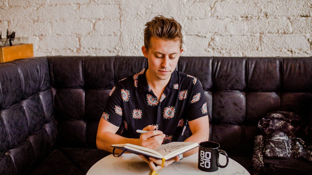 A masculine-appearing person with short hair and a patterned button-up sits in a leather booth at a cafe, looking down at an open notebook with a coffee cup and smartphone within reach. They appear lost in thought, pen in hand, as if contemplating something to write.