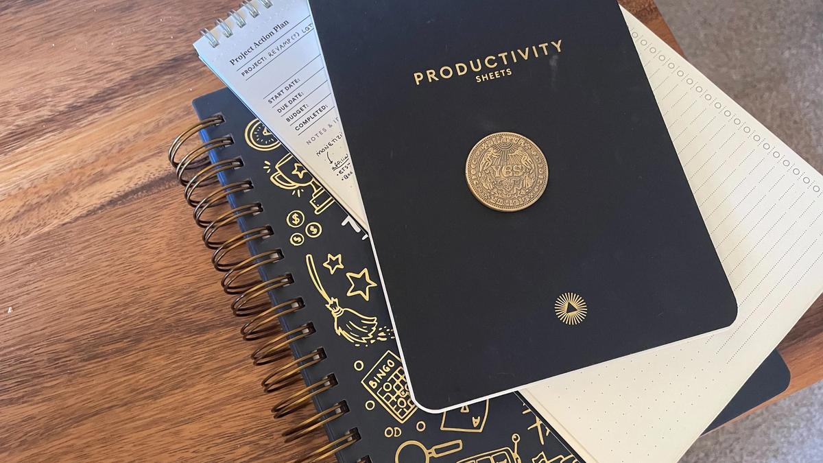 A stack of items on a desk, including a golden coin, two notepads, and a copy of Dani Donovan's "Anti-Planner" book peaking out from underneath.