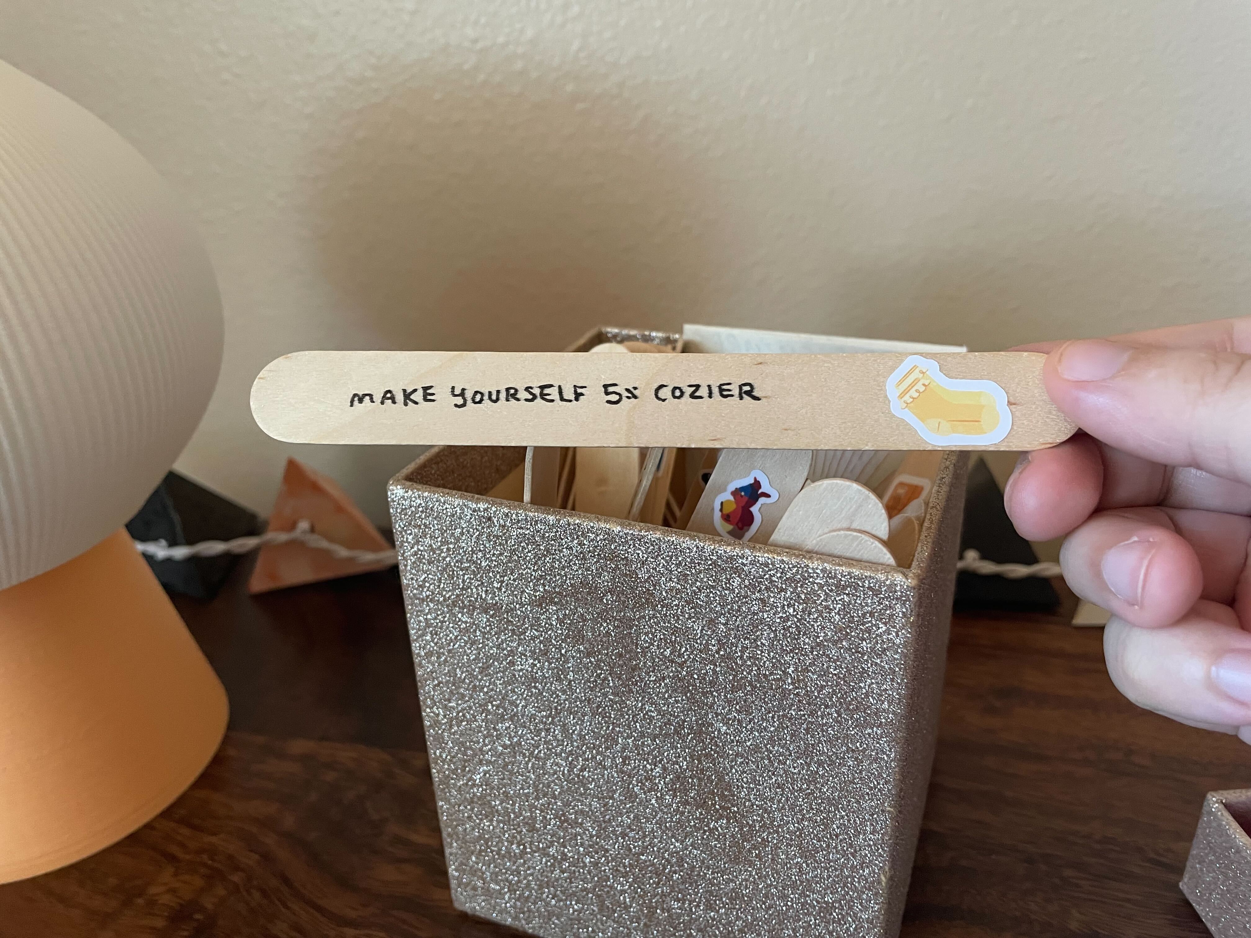 A sparkly gold box filled with popsicle sticks. A hand retrieves a popsicle stick that has writing on it: "Make yourself 5% cozier." It has a sticker next to the writing, featuring a pair of yellow socks.