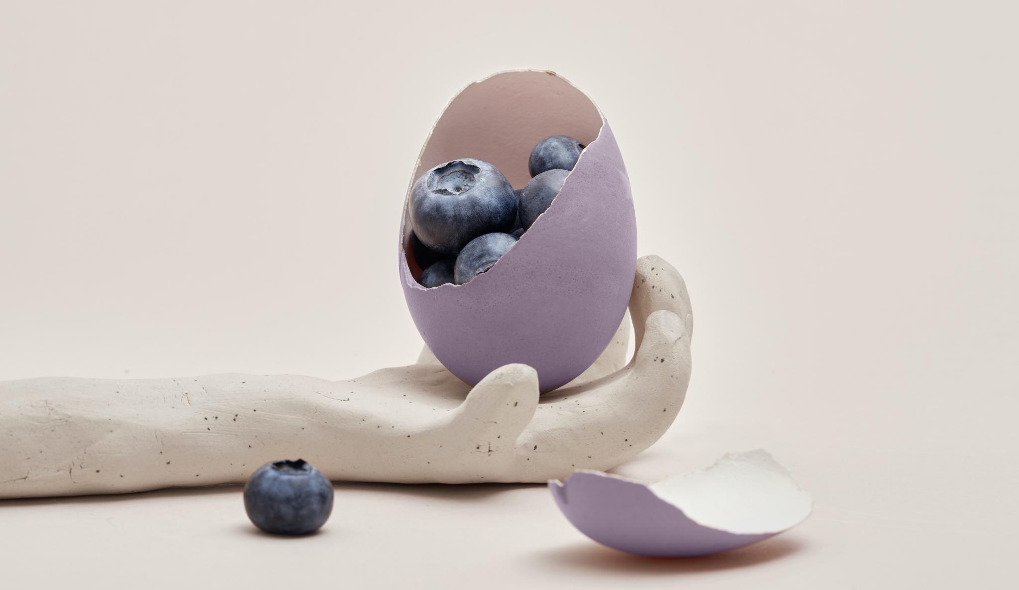 An outstretched hand made of clay, holding a purple egg whose shell has been cracked to reveal "superfood" blueberries inside. 