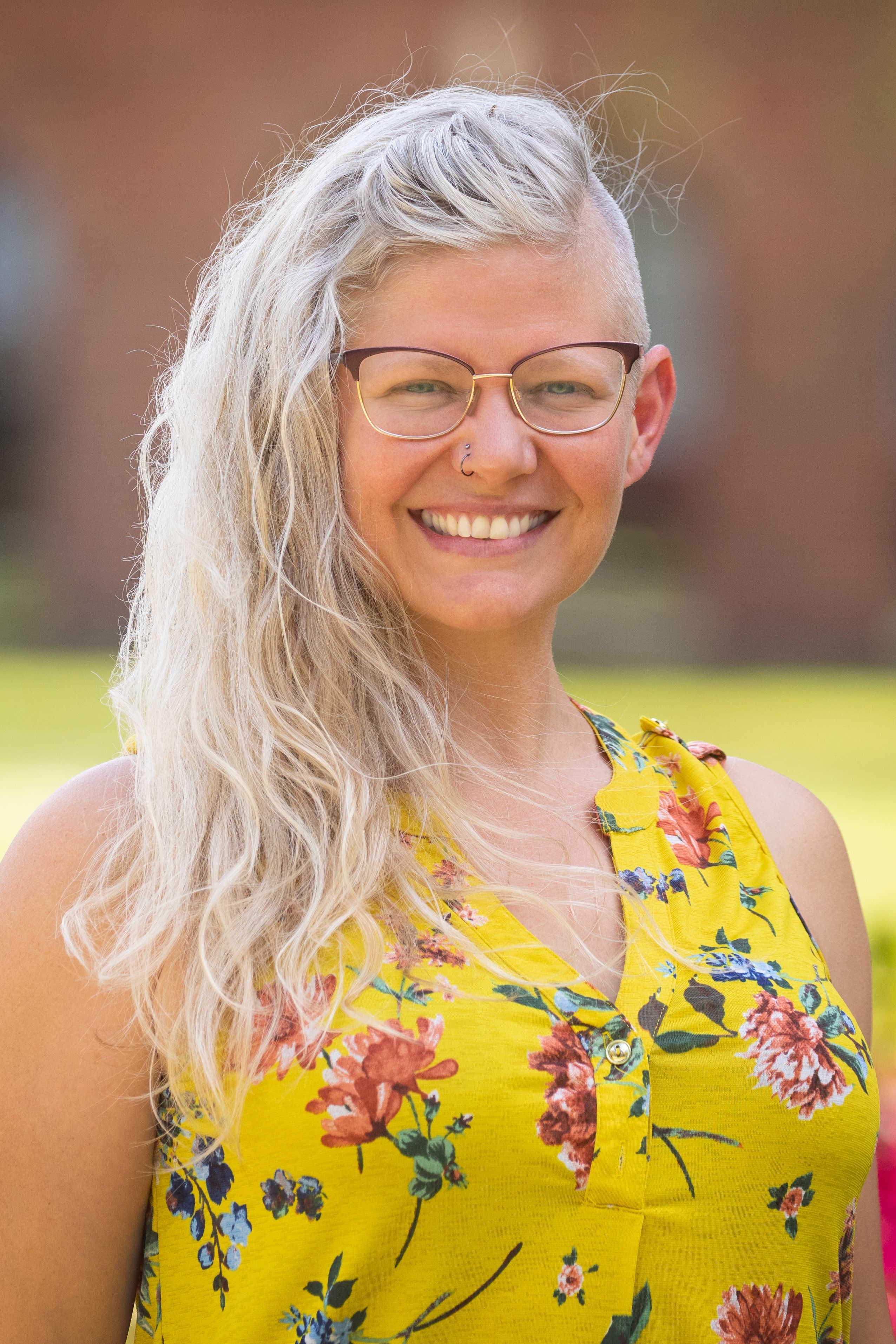 Dr. Emily K. Sandoz smiling in a bright yellow floral shirt with her silvery hair swept over one shoulder.