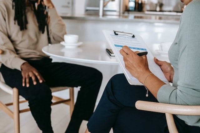 A therapist and their client seated at a café table in wishbone chairs, with the therapist taking notes on a clipboard.